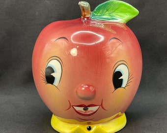 May include: A ceramic apple-shaped figurine with a smiling face, large eyes, and a yellow collar. The apple is primarily red with a green leaf and a brown stem. The figurine has a vintage, cartoonish design.