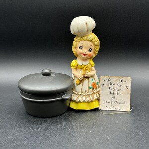 May include: A vintage ceramic figurine of a smiling chef in a white hat and yellow dress, holding a wooden spoon. Beside it is a small black pot with a lid, and a tag that reads "Handy Kitchen Hints A Josef Original."