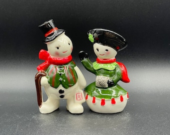May include: Two ceramic snowman figurines. One snowman wears a black top hat, red scarf, and green vest with a cane. The other snowman wears a black hat, green dress, and red scarf. Both have white bodies and are smiling.