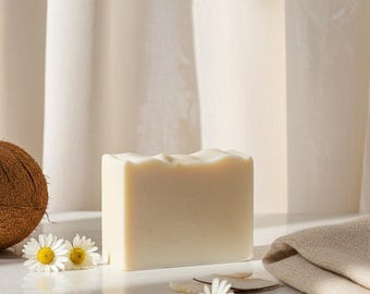 May include: A rectangular bar of off-white soap positioned next to a brown coconut, two white daisy flowers with yellow centers, and a folded beige fabric cloth. The soap has a smooth, solid appearance with a slightly uneven top edge.