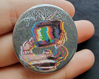 May include: A round button with a glitchy, pixelated image of a person with a television for a head. The text "STAND BY" is written in white on a black background.