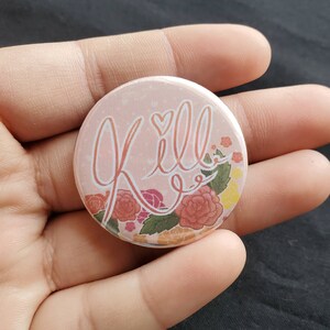 May include: A round, light pink button with the word "Kill" in a cursive font, with a heart above the double "l"s. The button is decorated with floral elements in shades of red, pink, and yellow.