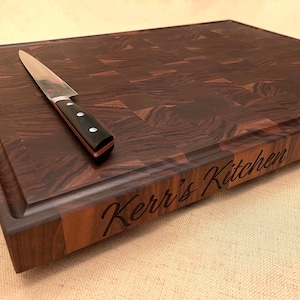May include: A dark wood butcher block cutting board with a knife resting on top. The board has a checkerboard pattern and a juice groove. The words "Kerr's Kitchen" are engraved on the front.