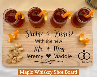 May include: A wooden shot board with the text "Shots & Kisses With the new Mr. & Mrs. Jeremy Maddie est. 10.14.22" and a heart symbol. The board has four shot glasses with orange peels and cashews.