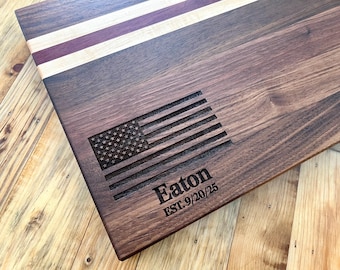 May include: A rectangular wooden cutting board with an engraved American flag design. The board features a dark brown wood grain with stripes of light wood and purple. The name "Eaton" and the date "EST. 9/20/25" are also engraved.