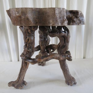 May include: A handcrafted wooden side table with a natural, rustic aesthetic. The tabletop is a thick, irregular wooden slab, supported by a base of intertwined branches and roots. The wood's natural tones vary from light to dark brown, highlighting the organic texture and unique design.