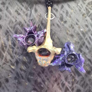 May include: A pendant necklace featuring three bone-like structures suspended from a gold-toned metal collar. The central piece is natural beige with orange and blue speckles, while the two flanking pieces are painted a deep, vibrant purple.