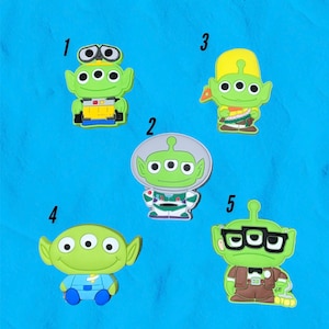 May include: Five green alien character figurines with different expressions and outfits. The aliens have three eyes and are wearing different colored clothing. The figurines are numbered 1 through 5.