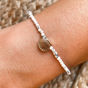 May include: A bracelet with white and black speckled beads and a gold seashell charm. The bracelet is strung on a flexible cord and is designed to be worn on the wrist. The bracelet is a delicate piece of jewellery.