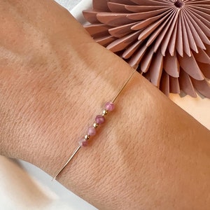 May include: A delicate bracelet featuring a thin metallic wire band adorned with three small, faceted pink gemstone beads separated by two tiny, smooth gold-toned spherical beads arranged in a linear pattern along the centre of the wire.