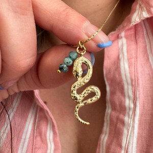 May include: A gold-coloured snake pendant necklace with a textured design. The snake is coiled and has a small cluster of blue-green beads. The necklace is held up by a person wearing a pink and white striped shirt.
