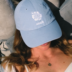 May include: A light blue baseball cap with a white hibiscus flower and the word "seak" embroidered on the front.