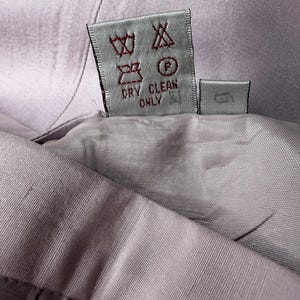 May include: Close-up of a lavender-colored garment with a care label. The label displays various symbols indicating washing instructions, including "DRY CLEAN ONLY". The fabric appears to be a smooth, woven material.