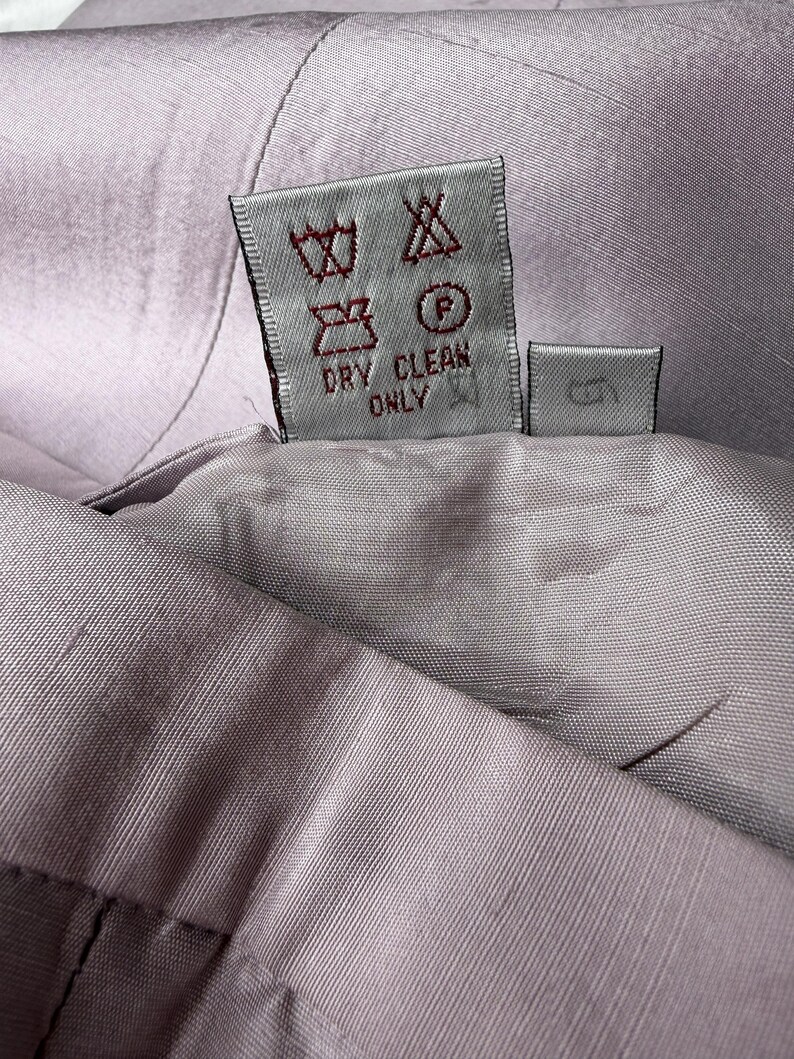 May include: Close-up of a lavender-colored garment with a care label. The label displays various symbols indicating washing instructions, including "DRY CLEAN ONLY". The fabric appears to be a smooth, woven material.