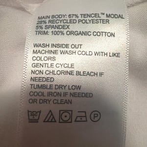 May include: A white fabric garment care label with black text listing the material composition as 67% Tencel Modal, 28% recycled polyester, 5% spandex, and 100% organic cotton trim, including washing instructions and five standard laundry care symbols.