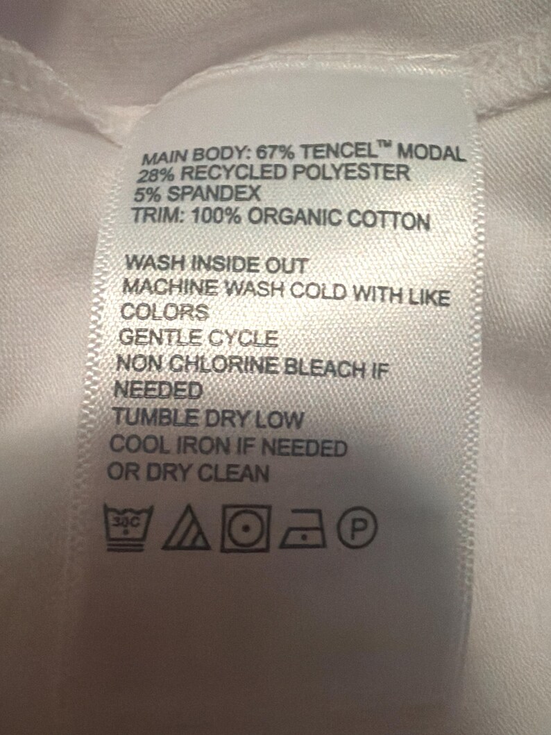 May include: A white fabric garment care label with black text listing the material composition as 67% Tencel Modal, 28% recycled polyester, 5% spandex, and 100% organic cotton trim, including washing instructions and five standard laundry care symbols.