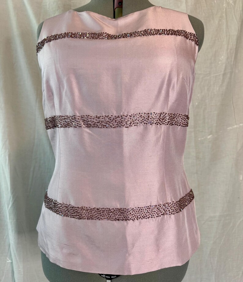 May include: A sleeveless, pale pink top with horizontal bands of small, dark beads. The top is made of a smooth, possibly silk-like fabric and has a fitted silhouette. The beaded bands add a touch of sparkle and detail to the garment.
