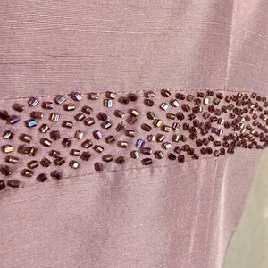 May include: Close-up of a lilac-colored fabric with a horizontal band of small, iridescent beads. The beads are a mix of brown and purple hues, adding a touch of sparkle to the garment. The fabric has a subtle sheen.