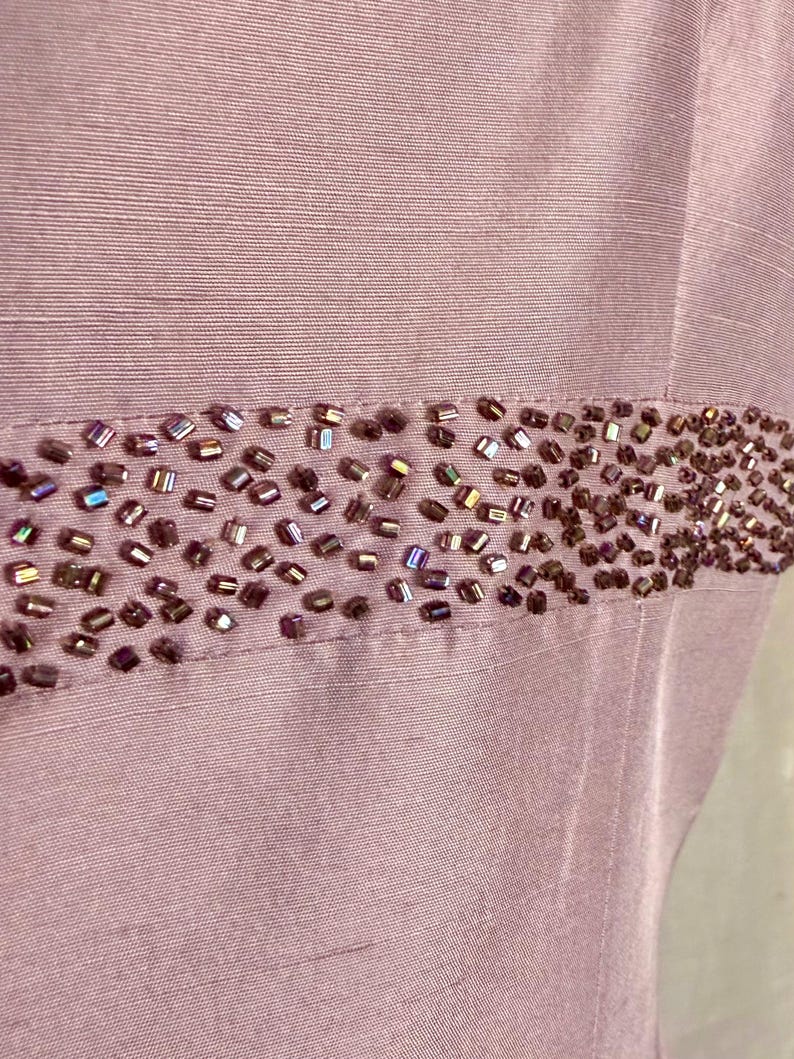 May include: Close-up of a lilac-colored fabric with a horizontal band of small, iridescent beads. The beads are a mix of brown and purple hues, adding a touch of sparkle to the garment. The fabric has a subtle sheen.