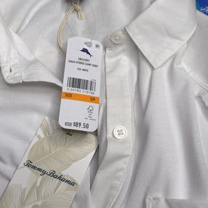May include: A white Tommy Bahama Kauai Hybrid camp shirt with a collar, a button-down front, and a chest pocket. The shirt includes a brand tag with the text Tommy Bahama and a product tag labeled Kauai Hybrid Camp Shirt, size S/P.