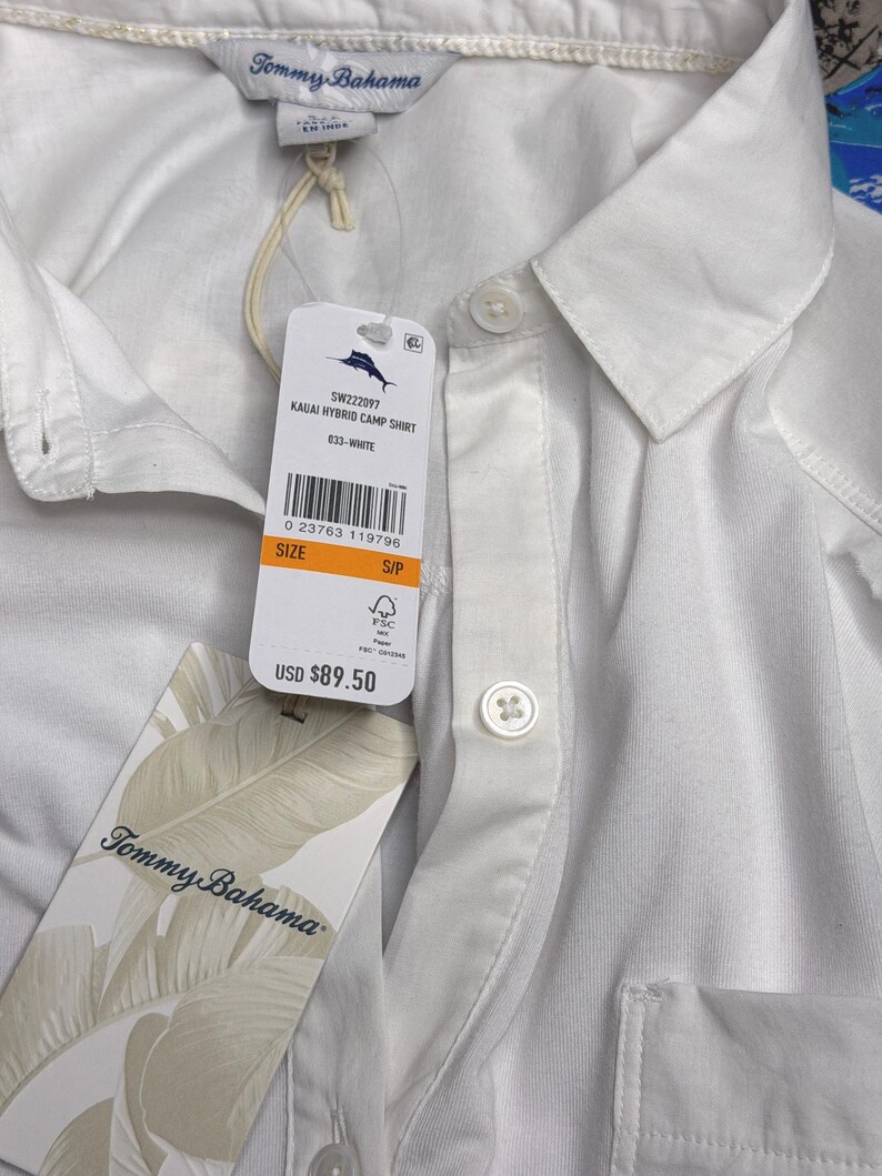 May include: A white Tommy Bahama Kauai Hybrid camp shirt with a collar, a button-down front, and a chest pocket. The shirt includes a brand tag with the text Tommy Bahama and a product tag labeled Kauai Hybrid Camp Shirt, size S/P.