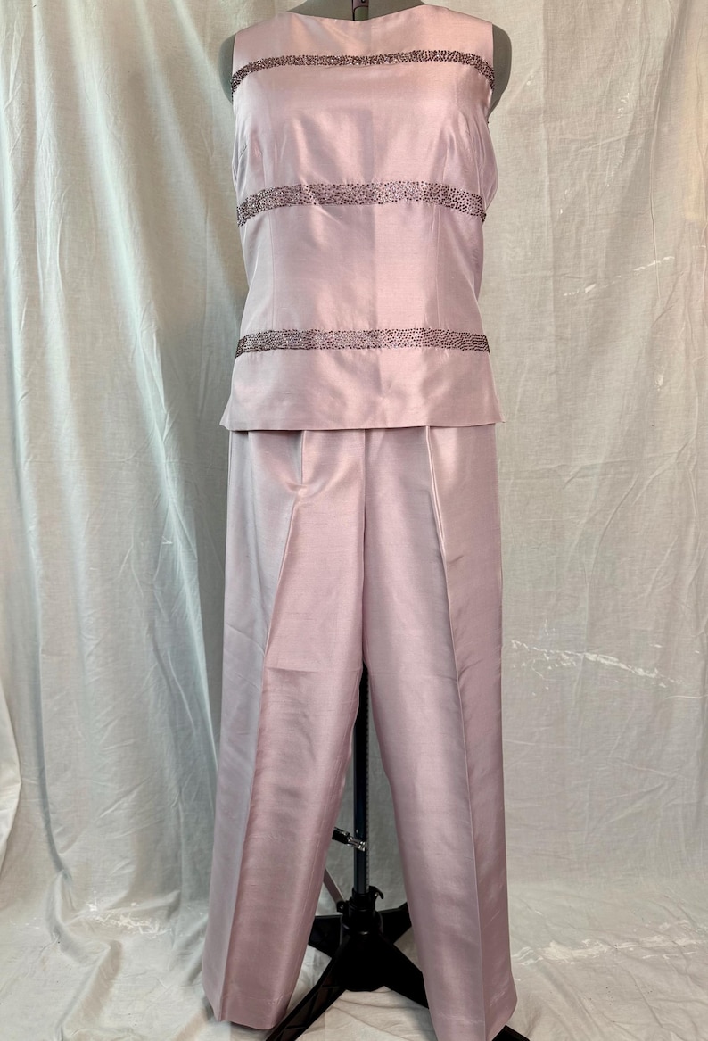 May include: A light pink two-piece formal outfit. The sleeveless top has three horizontal rows of dark beading. The matching pants are wide-legged. The outfit is displayed on a mannequin.