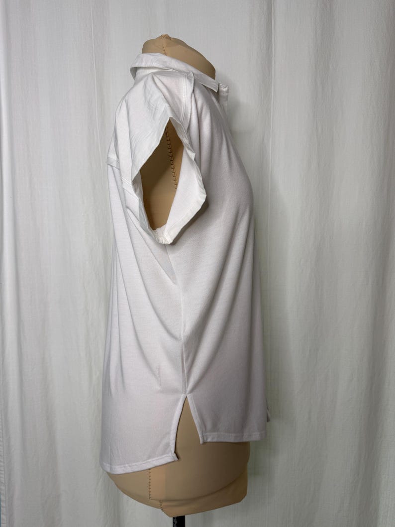 May include: A solid white short-sleeved polo shirt featuring a classic collar, a button-up placket, and side slits at the hem. The garment is displayed on a mannequin, showcasing a clean, plain design with a straight silhouette and hemline.