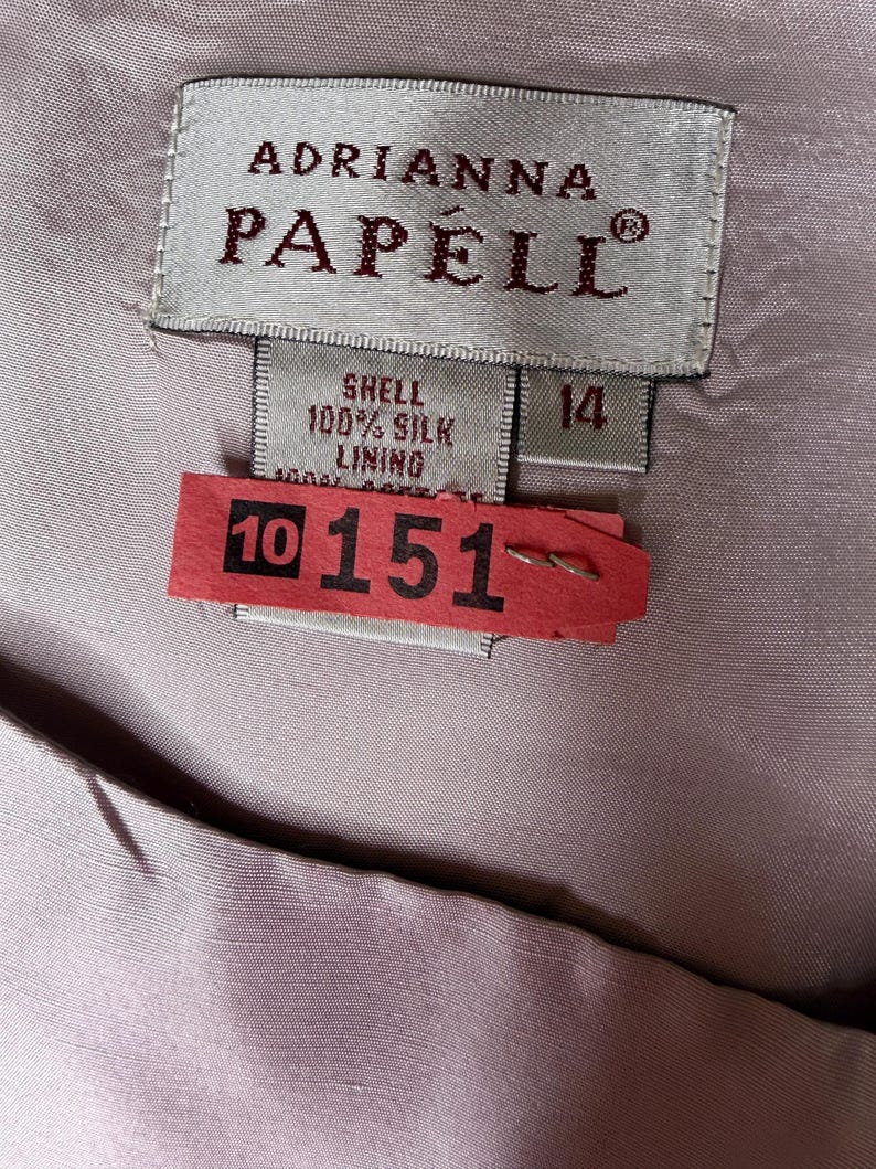May include: Close-up of a label from Adrianna Papell, size 14. The label reads "Shell 100% Silk Lining". A red tag with the numbers "10 151" is attached to the garment.