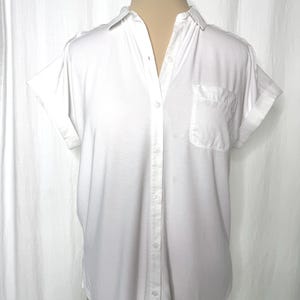 May include: A crisp white short-sleeved button-down shirt featuring a classic collar, a single chest pocket on the left side, and cuffed sleeves. The garment is constructed with a central vertical button placket and a rounded hemline.