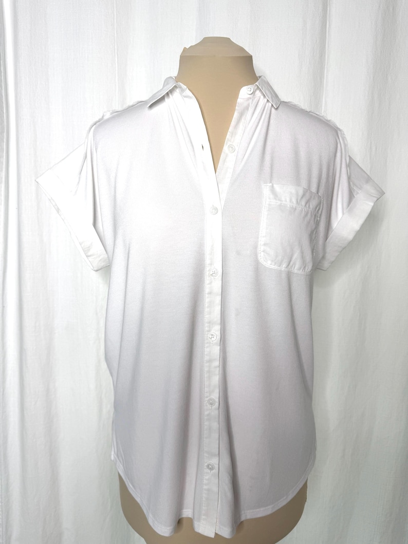May include: A crisp white short-sleeved button-down shirt featuring a classic collar, a single chest pocket on the left side, and cuffed sleeves. The garment is constructed with a central vertical button placket and a rounded hemline.