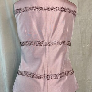 May include: A sleeveless, light pink top with horizontal bands of small, dark embellishments. The back of the top is visible, showing a zipper closure and a tailored fit. The fabric has a subtle sheen, suggesting a silk or satin material.