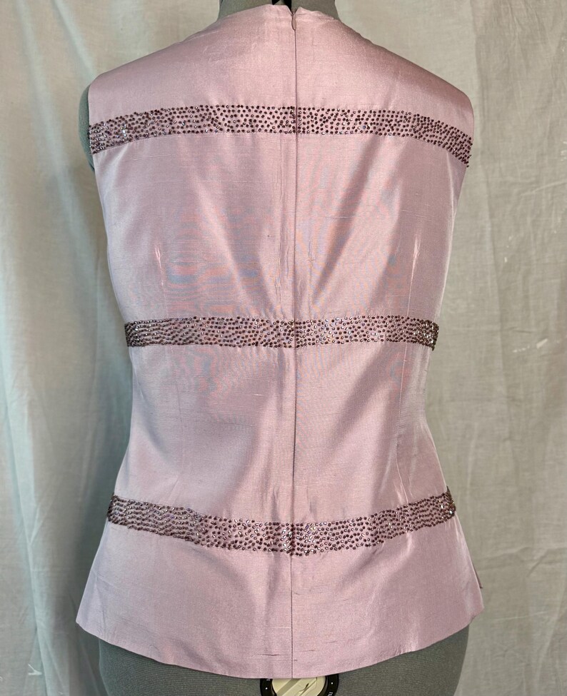 May include: A sleeveless, light pink top with horizontal bands of small, dark embellishments. The back of the top is visible, showing a zipper closure and a tailored fit. The fabric has a subtle sheen, suggesting a silk or satin material.