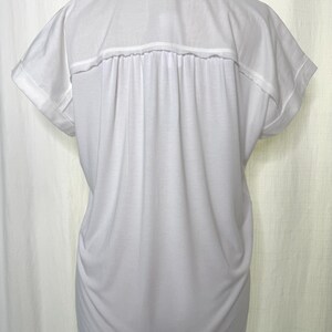 May include: A solid white short-sleeved shirt featuring a classic collar, a horizontal back yoke, and vertical pleats extending from the yoke down the back. The sleeves have folded cuffs, and the garment has a relaxed, loose-fitting silhouette.