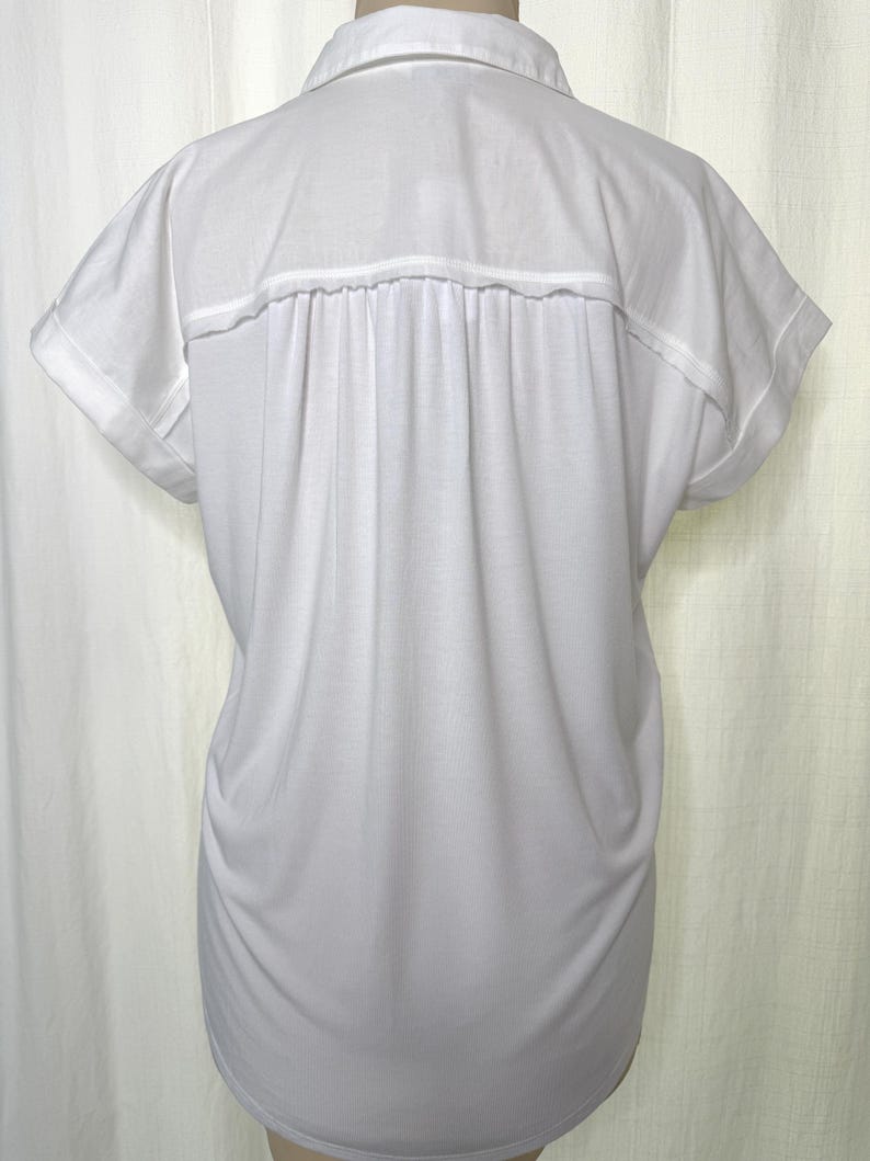 May include: A solid white short-sleeved shirt featuring a classic collar, a horizontal back yoke, and vertical pleats extending from the yoke down the back. The sleeves have folded cuffs, and the garment has a relaxed, loose-fitting silhouette.