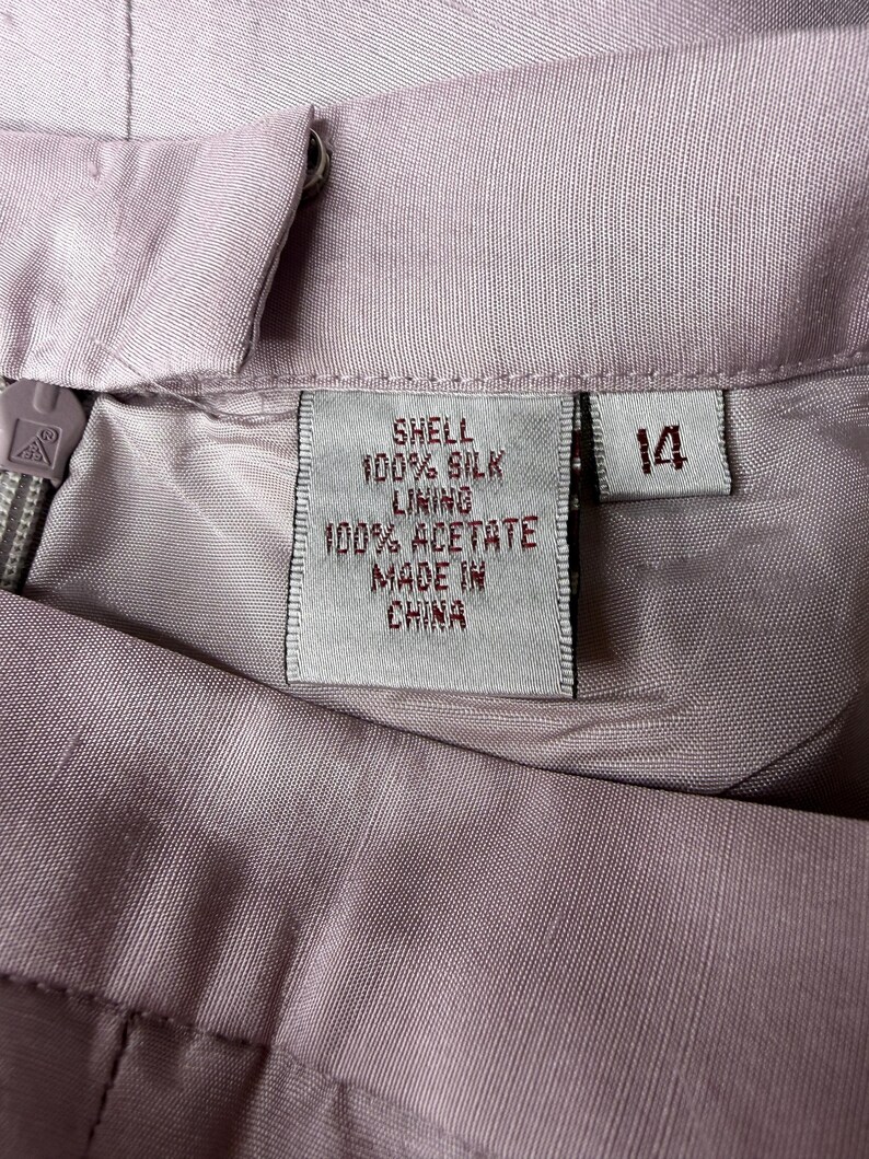 May include: Close-up of a light purple garment with a label indicating it's made of 100% silk shell and 100% acetate lining. The label also states "Made in China" and the size is 14.