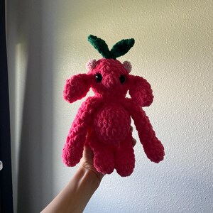 May include: A handmade, bright pink crochet creature with large, floppy ears and small, black eyes. The plush toy has green leaves and small pink accents on its head. The creature is held up against a plain background.