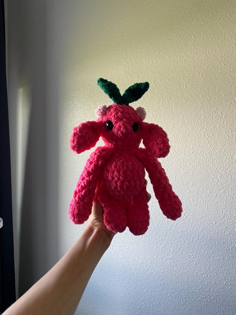 May include: A handmade, bright pink crochet creature with large, floppy ears and small, black eyes. The plush toy has green leaves and small pink accents on its head. The creature is held up against a plain background.