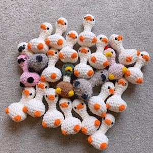 Op de afbeelding: Een verzameling handgemaakte gehaakte ganzen in verschillende kleuren. De ganzen zijn voornamelijk wit met oranje snavels en voeten. Sommige ganzen hebben roze, grijze of bruine lichamen. Het speelgoed is gerangschikt op een grijze ondergrond.