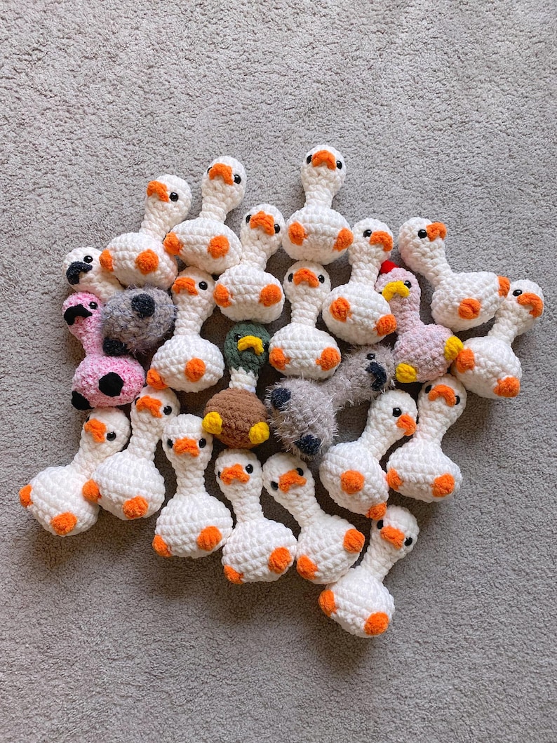 Op de afbeelding: Een verzameling handgemaakte gehaakte ganzen in verschillende kleuren. De ganzen zijn voornamelijk wit met oranje snavels en voeten. Sommige ganzen hebben roze, grijze of bruine lichamen. Het speelgoed is gerangschikt op een grijze ondergrond.