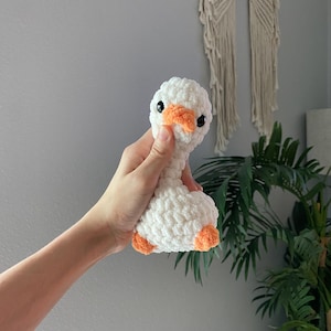 Op de afbeelding: Een handgemaakte witte gehaakte gans met een oranje snavel en poten. Het pluche speelgoed heeft zwarte veiligheidsoogjes en een zachte textuur. De gans wordt in de hand van een persoon gehouden.