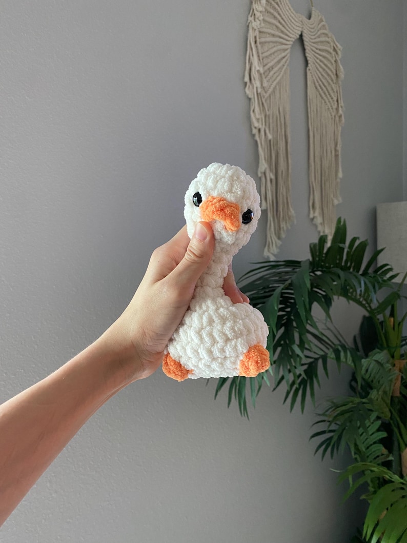 Op de afbeelding: Een handgemaakte witte gehaakte gans met een oranje snavel en poten. Het pluche speelgoed heeft zwarte veiligheidsoogjes en een zachte textuur. De gans wordt in de hand van een persoon gehouden.