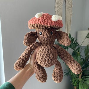 May include: A handmade brown crochet mushroom plushie with a coral-colored cap and white spots. The mushroom has a smiling face and outstretched arms and legs. The plushie is made of soft, textured yarn.