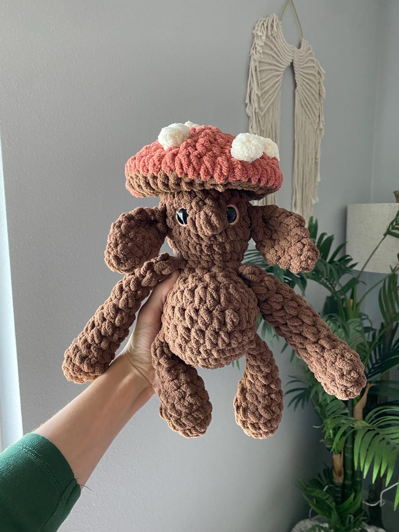 May include: A handmade brown crochet mushroom plushie with a coral-colored cap and white spots. The mushroom has a smiling face and outstretched arms and legs. The plushie is made of soft, textured yarn.