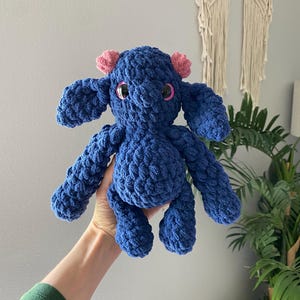 May include: A handmade, blue crochet stuffed animal with pink ear accents and large, round, black eyes. The plush toy has a textured, chunky appearance and is held up in front of a neutral background.