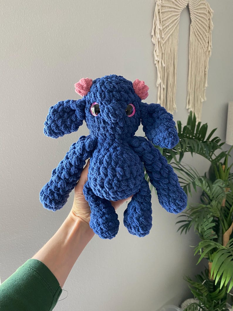 May include: A handmade, blue crochet stuffed animal with pink ear accents and large, round, black eyes. The plush toy has a textured, chunky appearance and is held up in front of a neutral background.