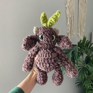 May include: A handmade, plush toy in shades of brown and green. The stuffed animal has a textured, fuzzy body, with green leaf-shaped accents on its head. It has small, round eyes and a cute, friendly appearance.