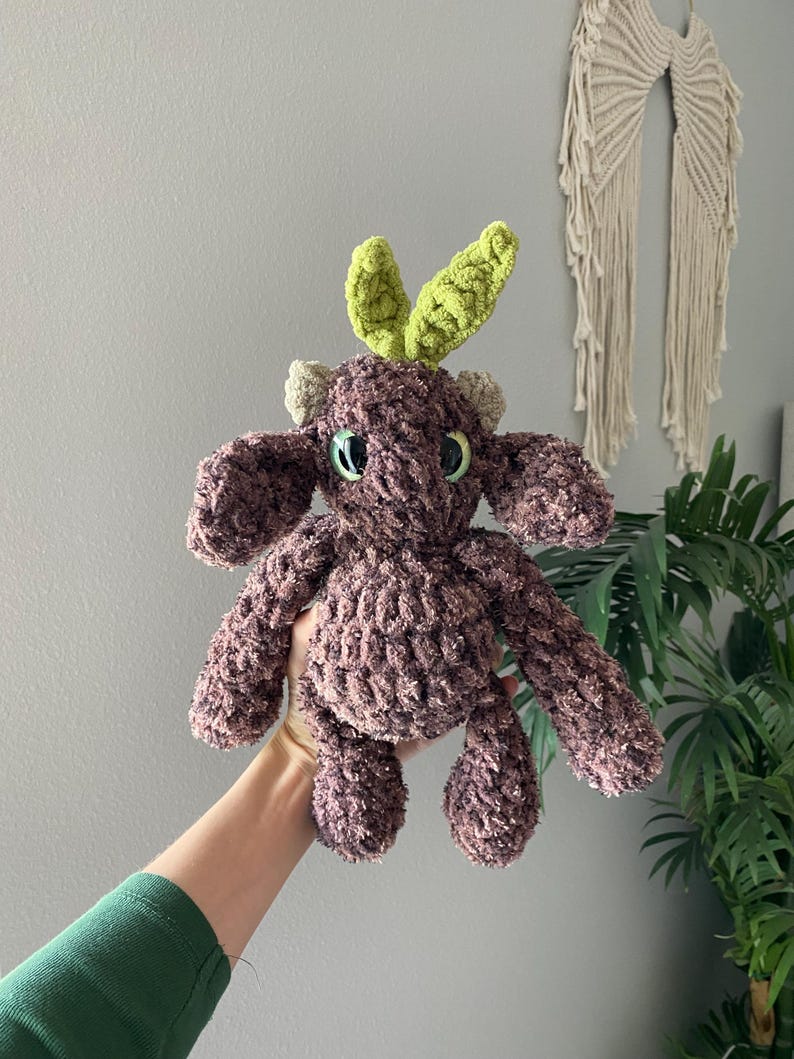 May include: A handmade, plush toy in shades of brown and green. The stuffed animal has a textured, fuzzy body, with green leaf-shaped accents on its head. It has small, round eyes and a cute, friendly appearance.