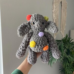 May include: A handmade gray crochet elephant plush toy with colorful pom-poms. The elephant has large ears, small eyes, and pom-poms in red, green, yellow, orange, and purple. The plush toy is soft and cuddly.