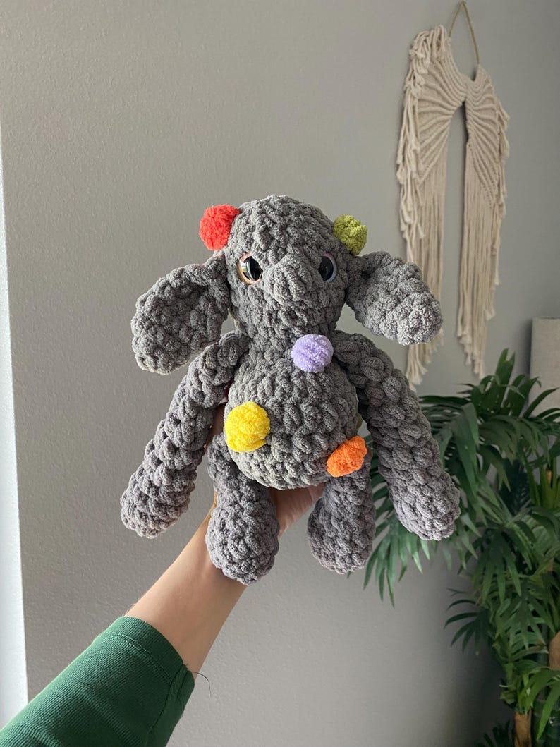 May include: A handmade gray crochet elephant plush toy with colorful pom-poms. The elephant has large ears, small eyes, and pom-poms in red, green, yellow, orange, and purple. The plush toy is soft and cuddly.
