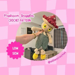 May include: A handmade crochet mushroom doll featuring a textured dark red cap with white spots, a beige body, yellow flower-shaped eyes, and sage green overalls. The doll has yellow feet and is labeled as a Mushroom Snuggler Crochet Pattern, Low Sew.